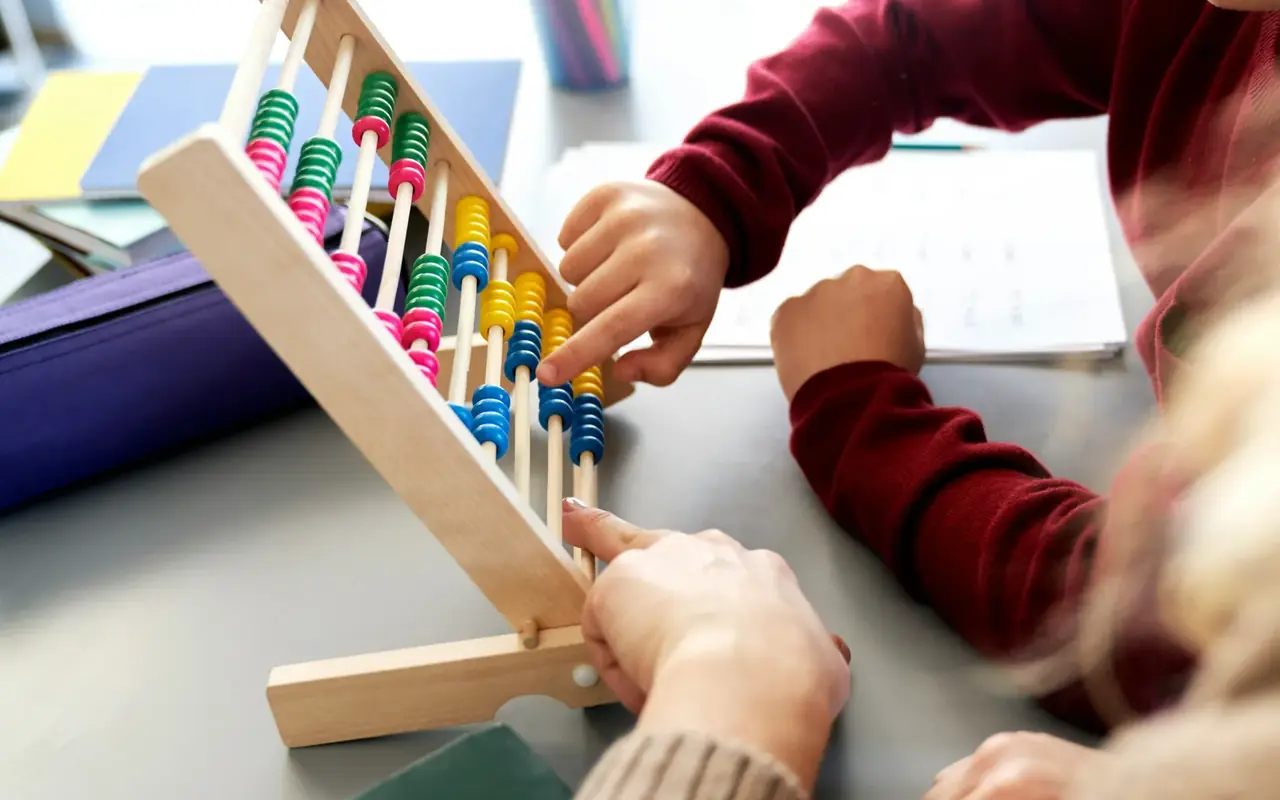 A teacher’s and a student’s hands working a wooden abacus together beside a paper worksheet